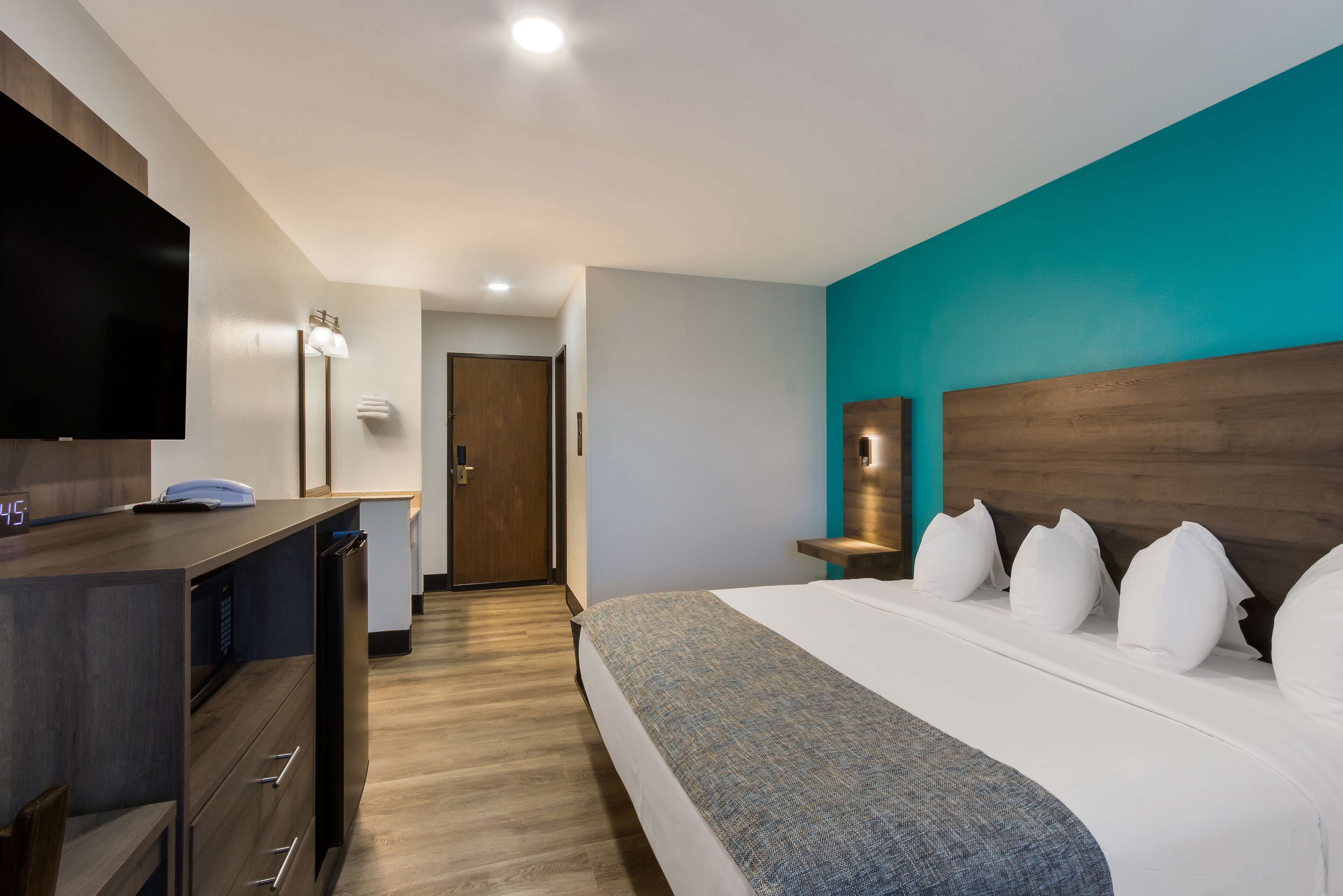 A hotel room with a bed, a desk with a telephone, a flat-screen TV, and a door leading to another room.
