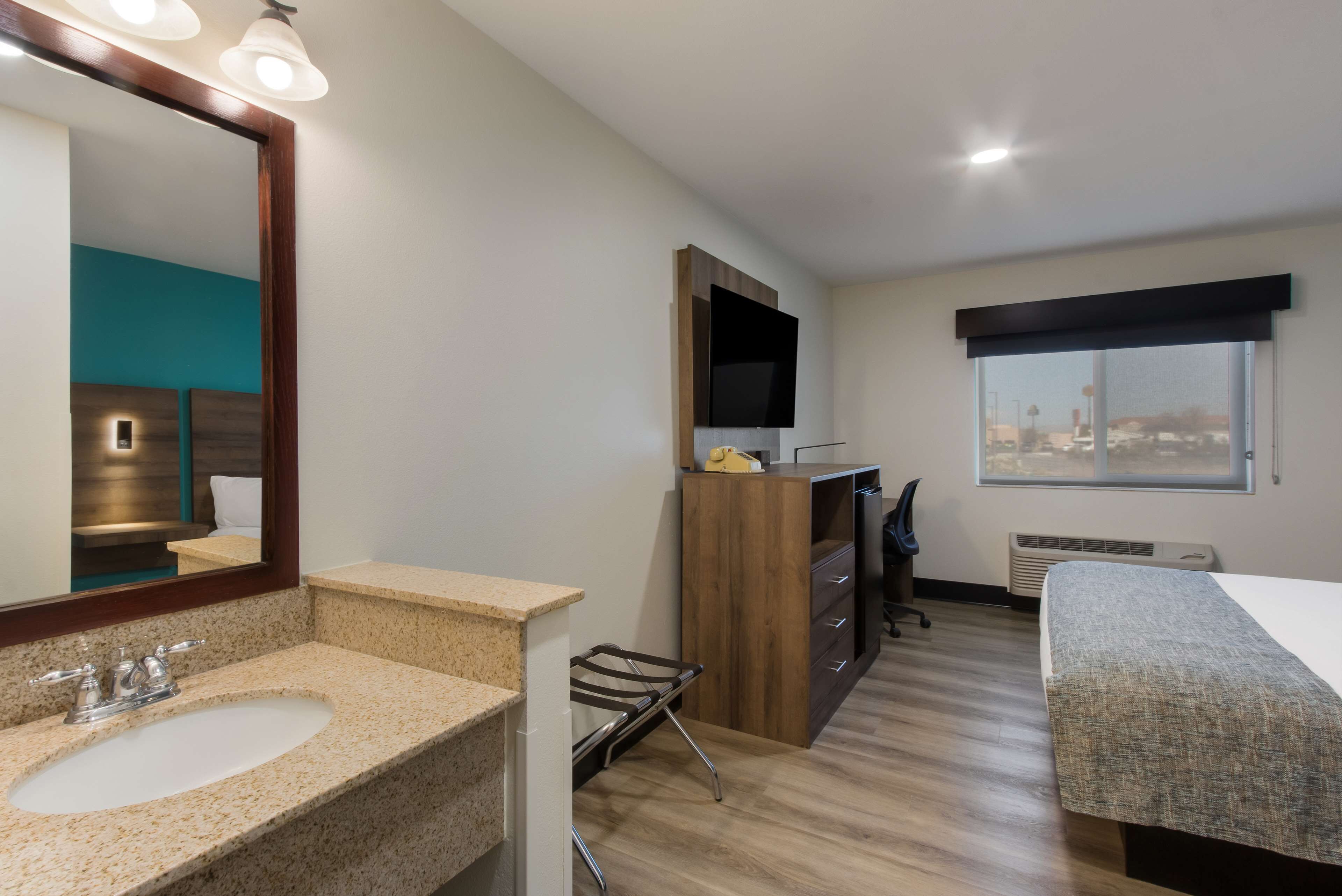 Habitación de hotel con un espejo grande, un lavamanos, un escritorio de madera, una silla, una cama y una ventana con vista a la ciudad.