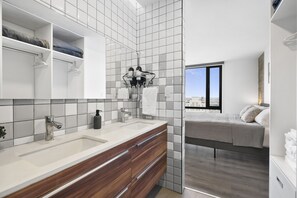 Ein modernes Badezimmer mit einem großen Spiegel, weißen Fliesen, einer Holzwaschbeckenbank und Blick auf ein Schlafzimmer mit Bett und Fenster.