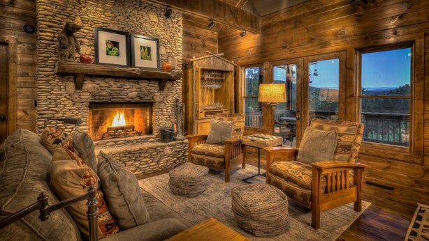 Interior acogedor de una cabaña con chimenea de piedra, muebles de madera y vistas al exterior.