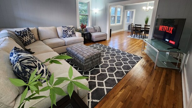 A modern living room with a sectional sofa, a glass coffee table, a black and white rug, a TV displaying 'NETFLIX', and a ceiling fan. - Spacious Hidden Gem (Utica)