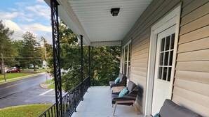 A covered porch with two wicker sofas and a small wooden table, overlooking a street with a car and trees. - Spacious Hidden Gem (Utica)