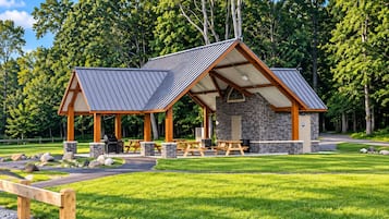 A rustic outdoor pavilion with picnic tables and a barbecue area, surrounded by a well-maintained lawn and trees.
