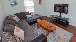 A living room with a gray sectional sofa, a wooden coffee table, a flat-screen TV, and a window with blinds. - Endless Summer - Resort Area - Vibe District VB (Virginia Beach)