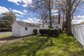 Une maison blanche à deux étages avec un garage, entourée d’une pelouse bien entretenue et d’un grand arbre.