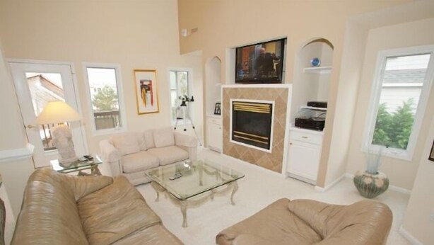 A living room with a beige sofa, a glass coffee table, a fireplace, and a television mounted on the wall. - A Wave From It All - Croatan VB (Virginia Beach)