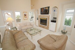 Ein Wohnzimmer mit einem beigen Sofa, einem Glastisch, einem Kamin und einem an der Wand montierten Fernseher.