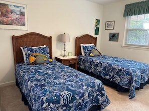 Two twin beds with matching blue floral bedspreads, a nightstand with a lamp, and a window with a view of greenery.