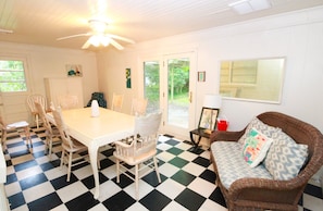A dining area with a checkered floor, a table, and four chairs. There is a wicker sofa with patterned cushions and a small table with a lamp.