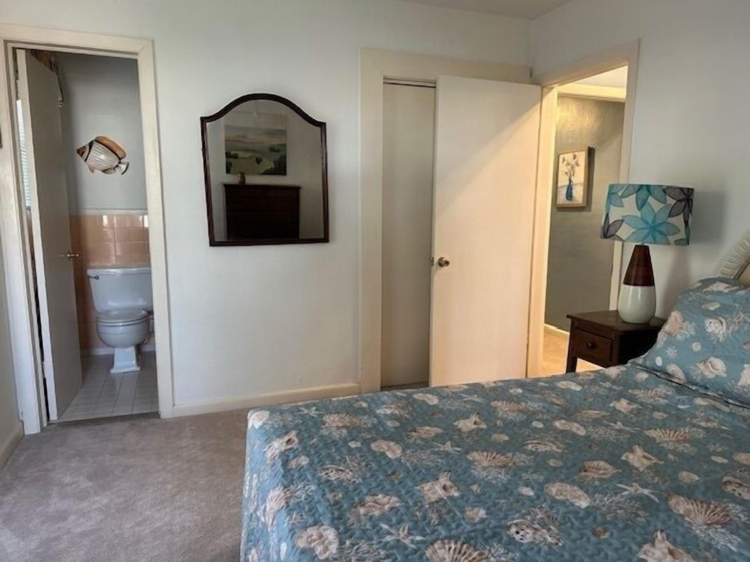 A bedroom with a bed, a nightstand, a lamp, a mirror, and a door leading to a bathroom.