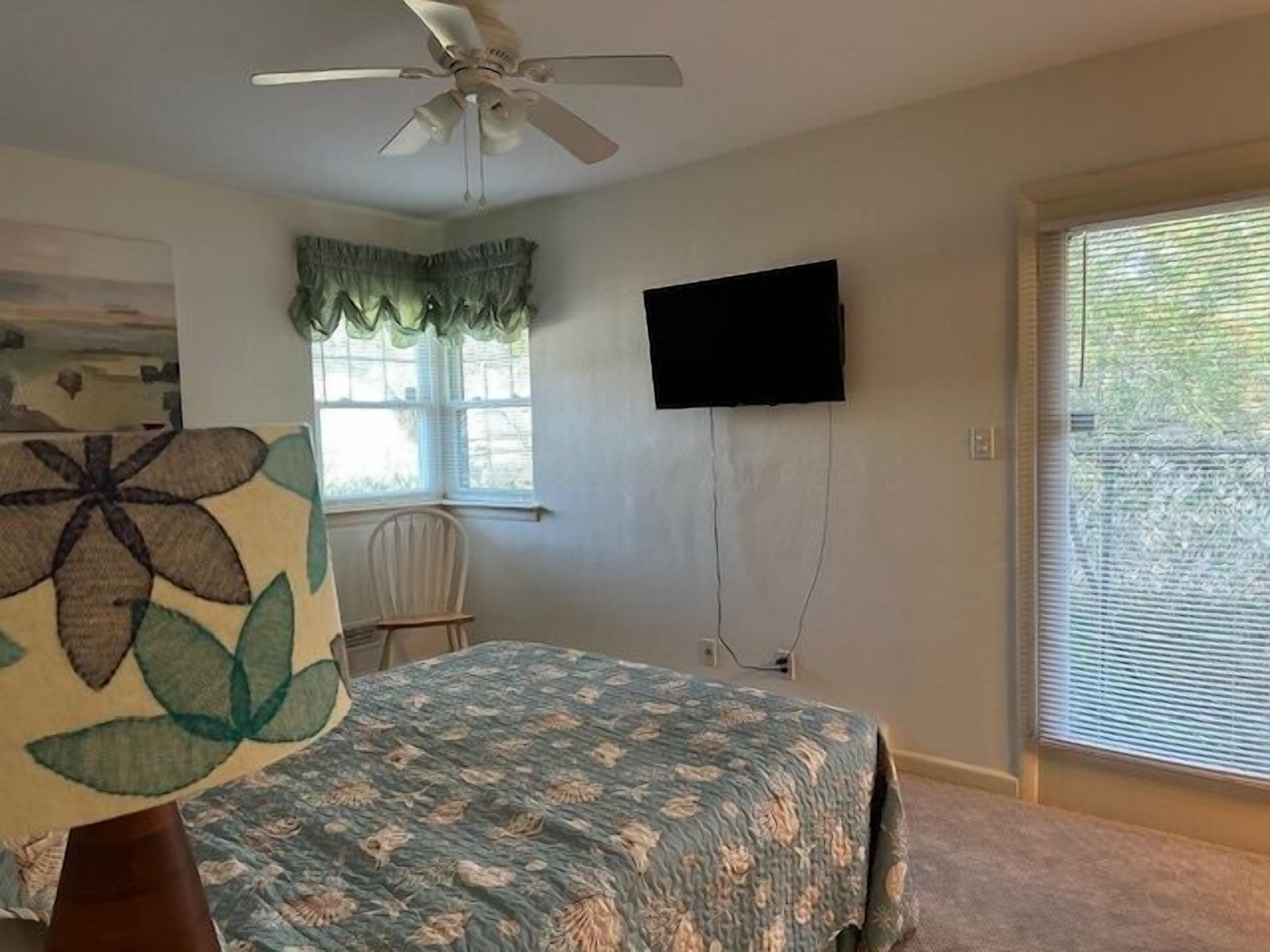 A bedroom with a bed, a ceiling fan, a TV mounted on the wall, and a window with blinds.
