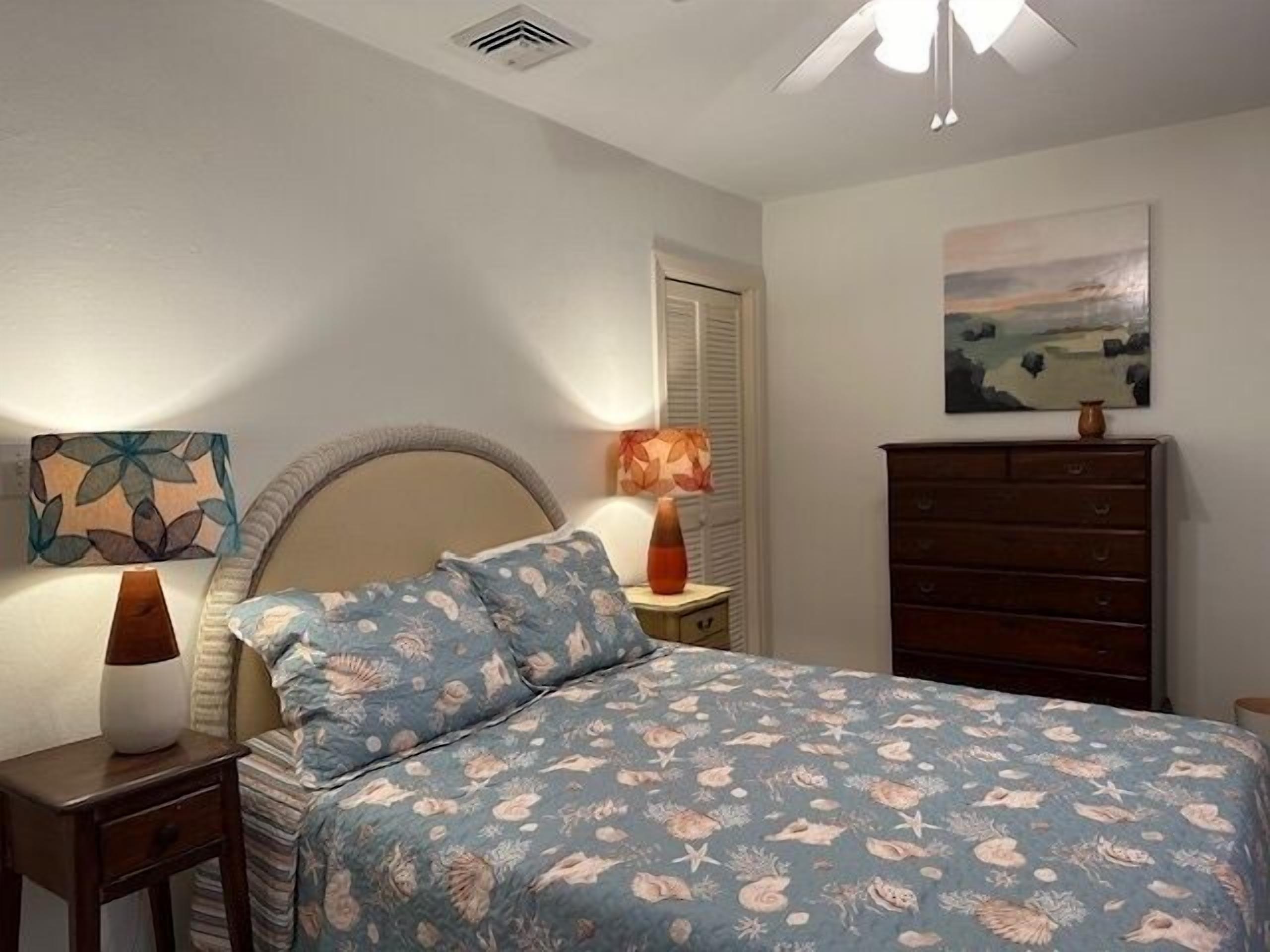 Un dormitorio con cama, mesitas de noche, cómoda, ventilador de techo, lámpara de pared y un cuadro.