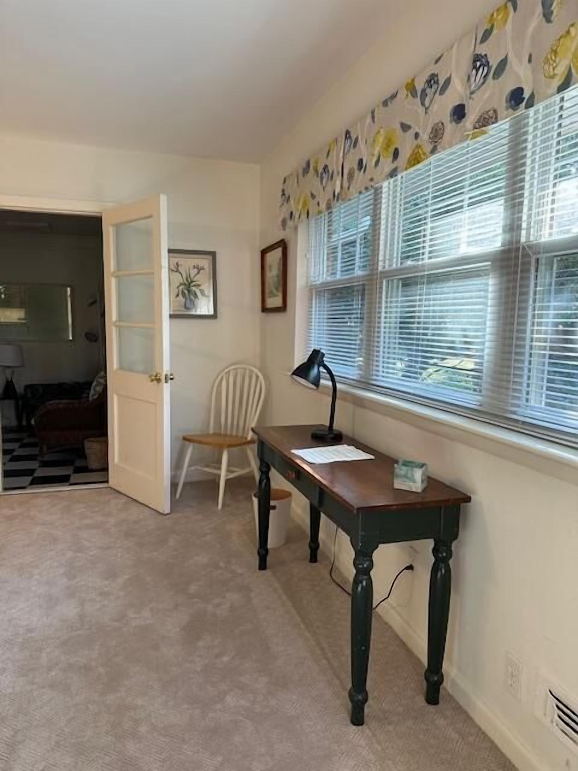 A room with a desk, chair, window with blinds, and a door leading to another room.