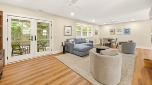 Un salon moderne avec un canapé d’angle, un fauteuil gris, un coin repas et un ventilateur de plafond.