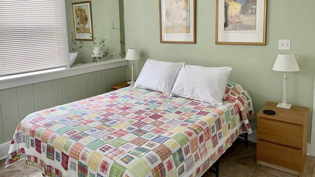 Une chambre à coucher avec un lit, deux tables de chevet, une fenêtre avec des stores et deux tableaux encadrés accrochés au mur.