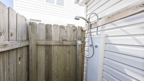 Une clôture en bois avec un portail en métal, une pomme de douche en métal fixée à la clôture et un mur extérieur blanc.