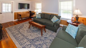 Ein Wohnzimmer mit einem grünen Sofa, einem Holztisch und einem gemusterten Teppich. Es gibt einen Esstisch mit Stühlen und ein an der Wand befestigtes Fernsehgerät.