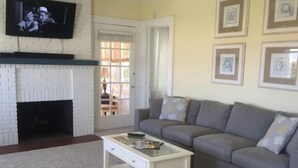 Een woonkamer met een wit bakstenen haardvuur, een grijze bank, een salontafel en een aan de muur bevestigde televisie.