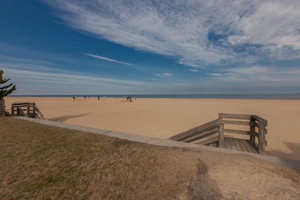 A beach with a wooden walkway leading to a sandy area, a grassy foreground, and a clear blue sky.