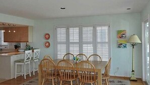 A dining area with a table and chairs, a kitchen area with a counter and cabinets, and a window with blinds. - Dolphin's Den - Oceanfront - North End VB (Virginia Beach)