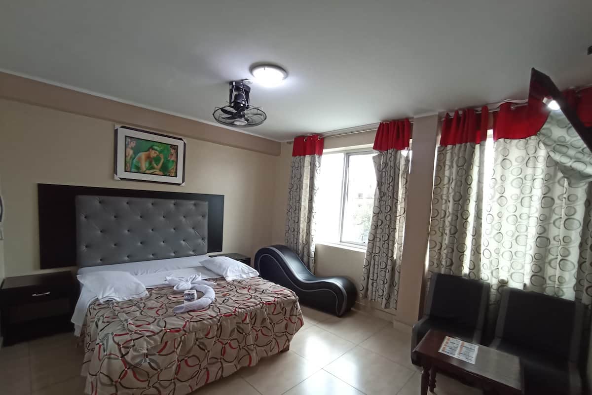 A hotel room with a bed, a chair, a standing fan, a window with curtains, and a framed picture on the wall.