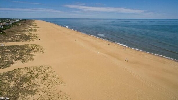 A wide sandy beach with gentle waves, a clear sky, and sparse vegetation. - Turtle Cove - North End VB (Virginia Beach)