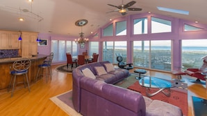 Un vaste espace de vie comprenant un canapé violet, une table à manger et des chaises. Une grande fenêtre offre une vue sur la plage.