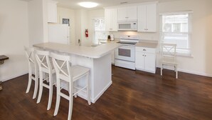 A modern kitchen with white cabinetry, a central island with bar stools, a microwave, oven, and a window with blinds. - North End Paradise - North End VB (Virginia Beach)