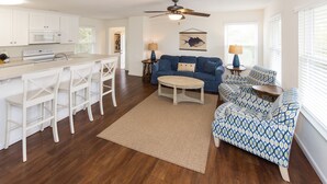 A modern kitchen with white cabinets, a microwave, and a stove. There is a dining area with white chairs and a table. A blue sofa and patterned armchair are in the living room, with a ceiling fan above. - North End Paradise - North End VB (Virginia Beach)