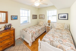 A bedroom with two single beds, a wooden dresser, a ceiling fan, and framed pictures on the walls.