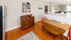 A kitchen with wooden cabinets, a dining table, and chairs. There is a refrigerator, a wooden cabinet, and a rug on the floor. - Conch Shell Cottage - North End VB (Virginia Beach)