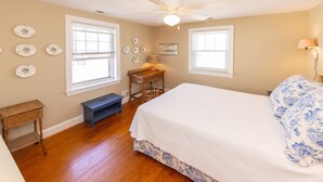 A bedroom with a large bed, two windows, a ceiling fan, and a desk with a lamp. - Conch Shell Cottage - North End VB (Virginia Beach)