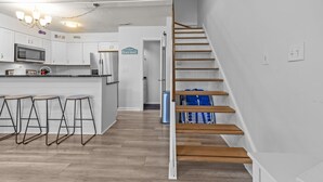 Moderni keittiö, jossa on valkoiset kaapit, keskellä sijaitseva saareke baarituoleineen, mikroaaltouuni ja jääkaappi. Puuportaat johtavat yläkertaan.