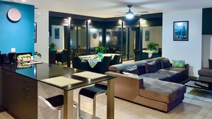 A modern living room with a sofa, a dining area with a table and chairs, and a kitchen area with a coffee machine and a clock on the wall.