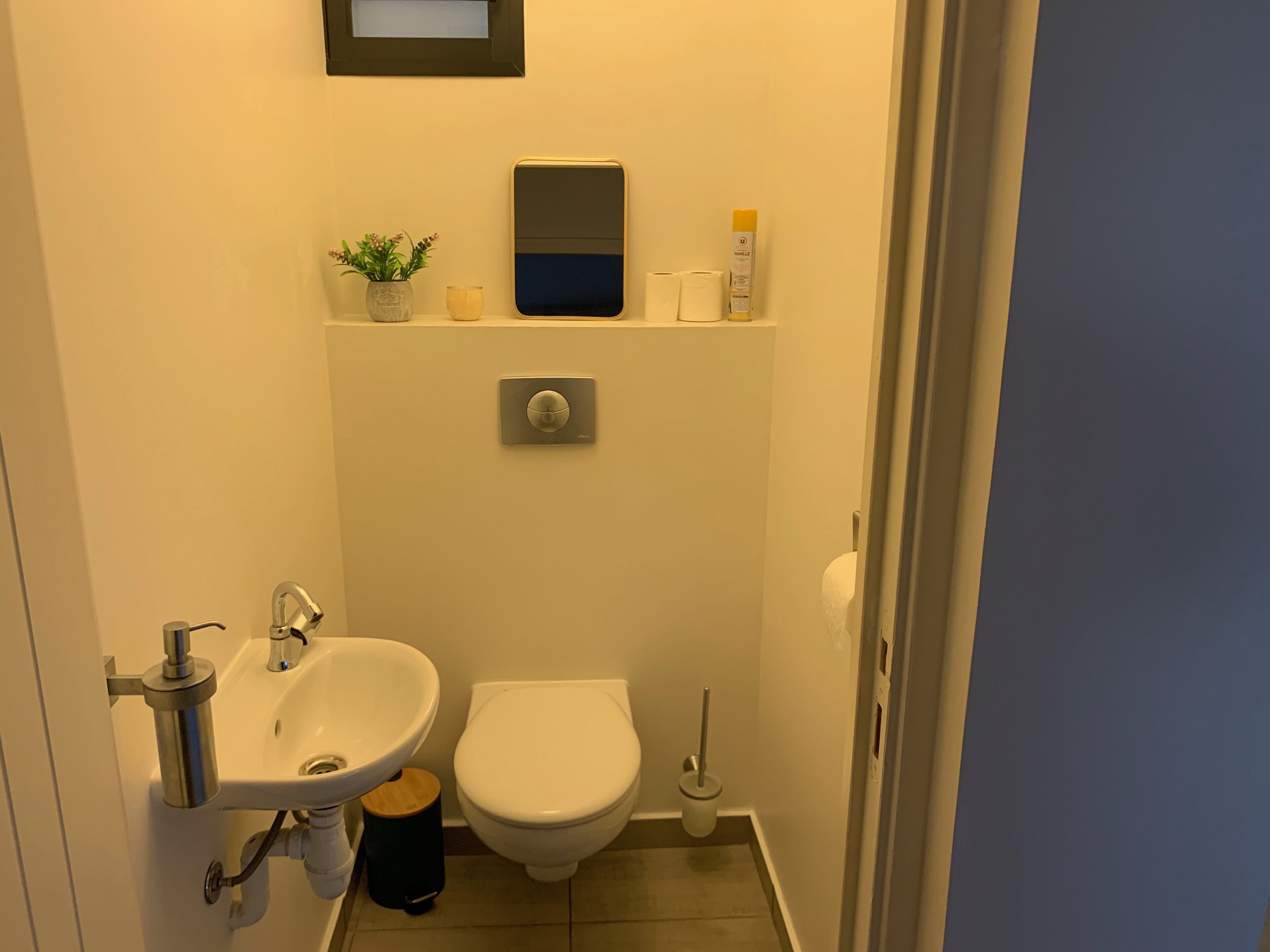A compact bathroom with a sink, toilet, and a small shelf with toiletries.
