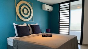 A bedroom with a bed, pillows, a blanket, an air conditioner, a lamp, and a view through a window.