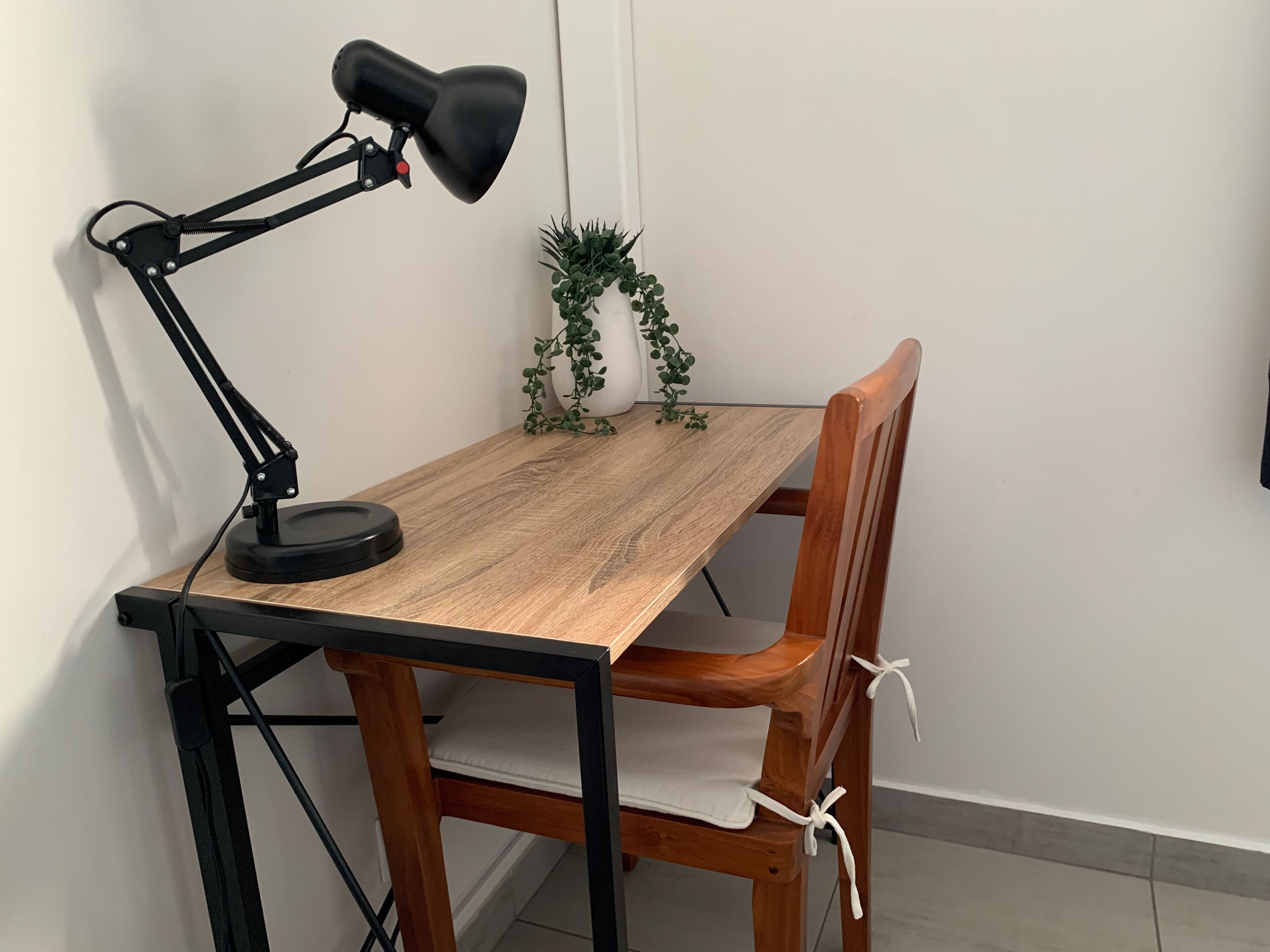Ein Holztisch mit einer schwarzen Schreibtischlampe, einem kleinen Topfpflanzen und einem Holzstuhl mit weißem Kissen.