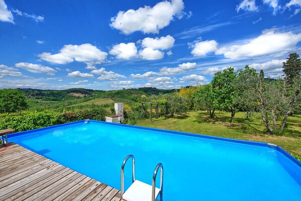 A swimming pool with a wooden deck, surrounded by lush greenery and a scenic view of hills.