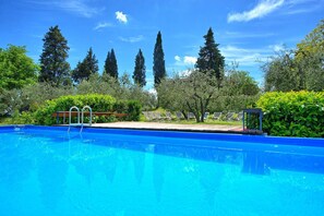 A swimming pool with a wooden deck and lounge chairs, surrounded by greenery and tall trees.