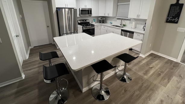 A modern kitchen with white cabinets, stainless steel appliances, a large island with bar stools, and a wooden floor. - Get Cozy in this newly renovated home by The Villages! (Lady Lake)