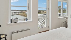 Quarto com três janelas amplas, uma cama, um radiador e uma pequena mesa com um livro e óculos.
