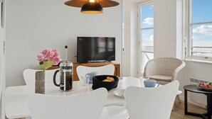 Uma área de refeições moderna com cadeiras brancas, uma cafeteira, uma televisão e uma cadeira de vime.