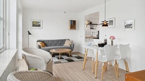 Uma sala de estar moderna com um sofá cinzento, cadeiras de vime, uma mesa de jantar com cadeiras brancas e uma área de cozinha visível através de uma abertura.