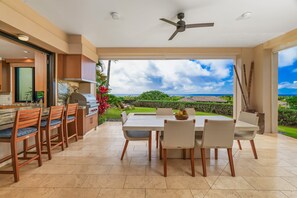 A modern kitchen with wooden cabinets and a large island, a dining area with a table and chairs, and a view of a lush garden and ocean. - Malu Makani by Alekona Kauai: 2 BR Luxury Oceanview Kukuiula Condo w private pool, AC, large lanai (Koloa)