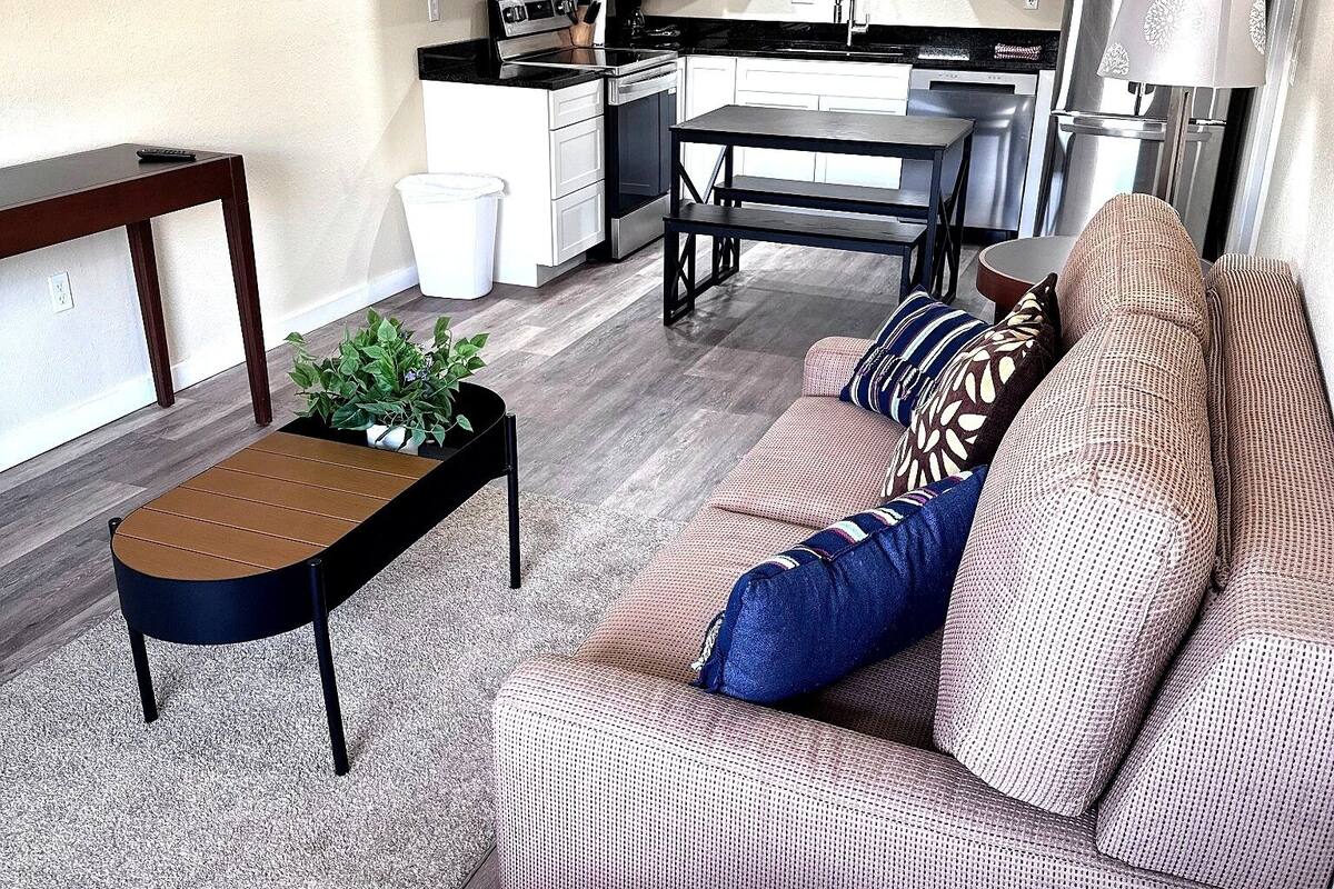 A living room with a sofa, a coffee table, and a dining table. There is a kitchen area with cabinets and appliances in the background.