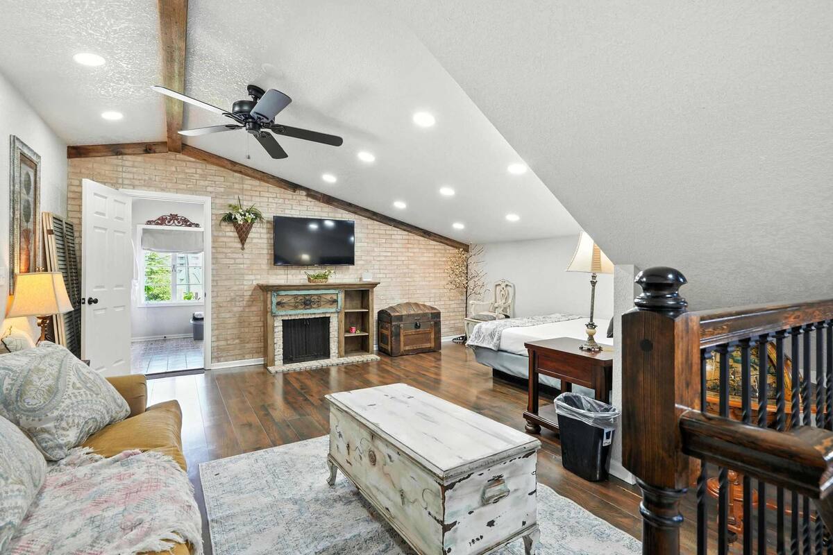 A loft-style room with a fireplace, a television, a ceiling fan, and a staircase.
