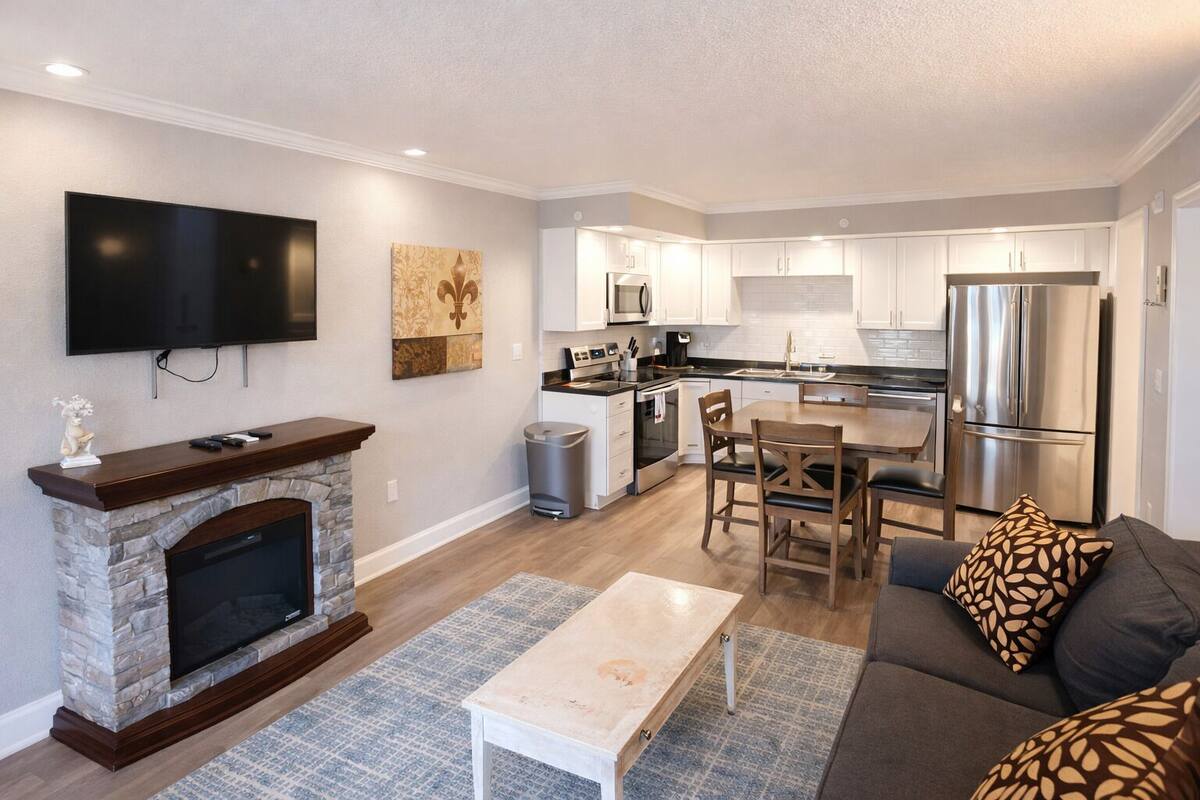A modern living room with a fireplace, a flat-screen TV, a dining area with a table and chairs, and a kitchen with stainless steel appliances.