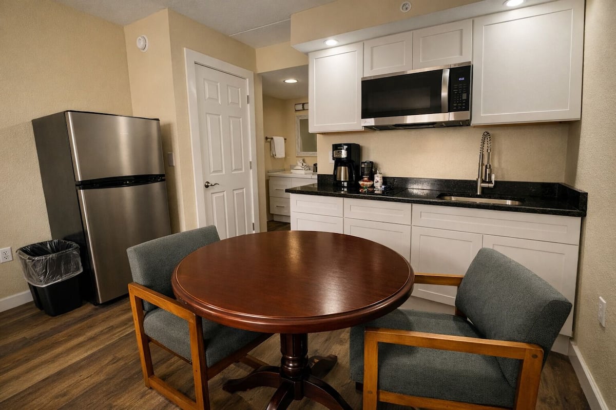 A compact kitchen with a stainless steel refrigerator, a microwave, a coffee maker, and a sink. There is a round wooden table with two chairs.