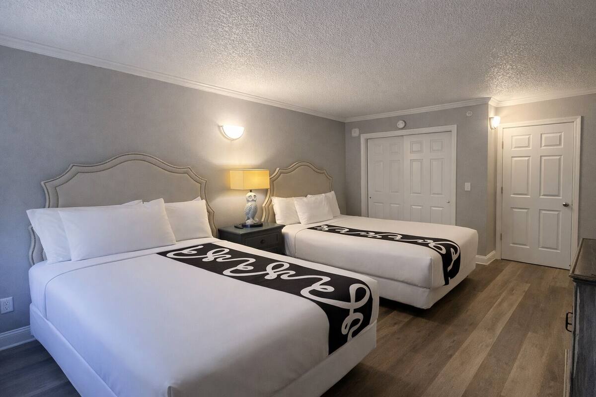 A hotel room with two beds, a nightstand with a lamp, and a door.