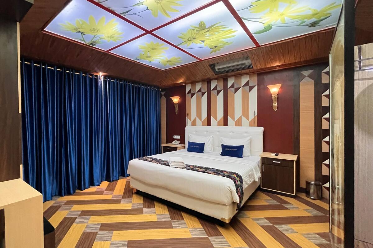 A hotel room with a large bed, a wooden floor, a blue curtain, and a ceiling with a floral design.